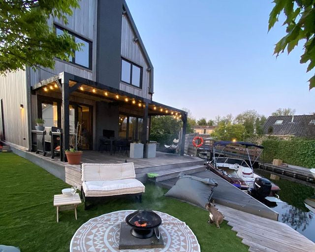 Moderne Douglas pergola met zwart gepoedercoate koppelstukken, bevestigd aan een gevel. De overkapping biedt sfeervolle verlichting en een stijlvolle uitbreiding van het terras. Perfect voor een luxe buitenruimte met bescherming tegen zon en regen. Geïntegreerd in een moderne tuin aan het water
