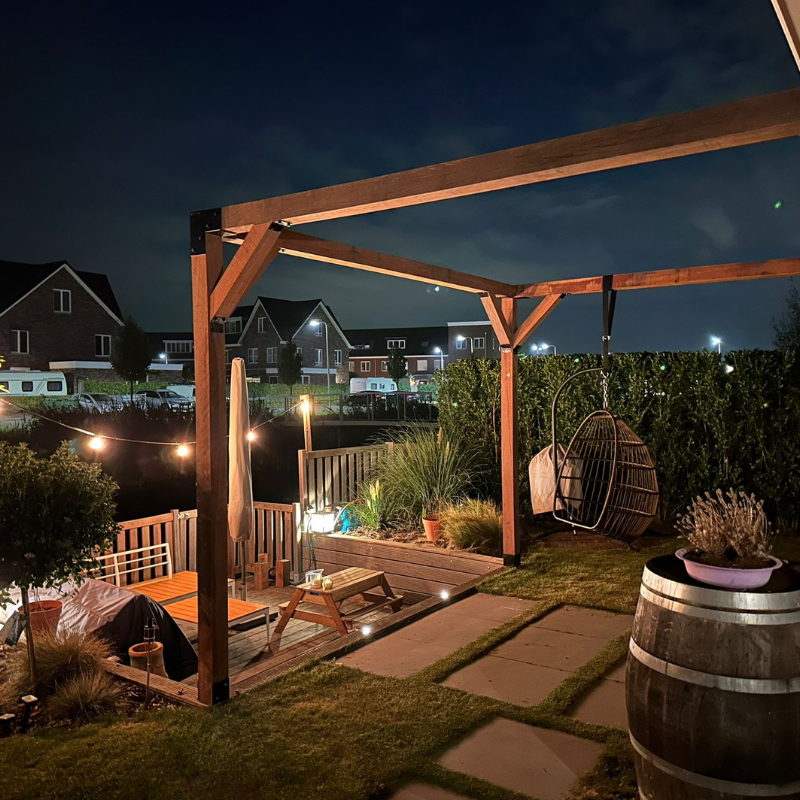 Houten pergola (12x12 cm) aan de muur gemonteerd, sfeervol verlicht in een tuin bij nacht. Douglas constructie met metalen koppelstukken voor een luxe buitenruimte