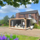 Houten muurpergola (15x15 cm) met wit harmonicadoek, gemonteerd aan een landelijke woning. Luxe terrasoverkapping van Douglas hout, perfect voor een sfeervolle buitenruimte